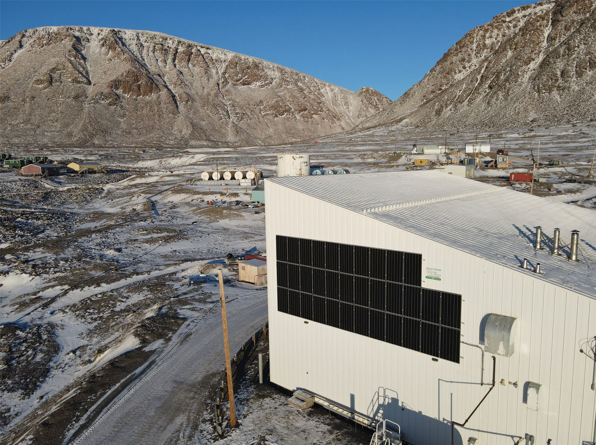 Wide shot of community hall with solar panels on the side and oil tanks in the distance beside mountains.