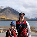 A mother and her child wearing traditional clothing from the killer whale clan, standing in front of a river.