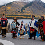Five dancers dressed in regalia stand in front of a body of water and snow-capped mountains.