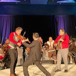 Fiddle and guitar players performing onstage, with Minister Vandal dancing to the left.