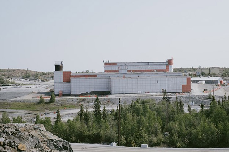 Un grand bâtiment industriel recouvert de plastique sur un chantier de construction avec des rochers et des arbres au premier plan.