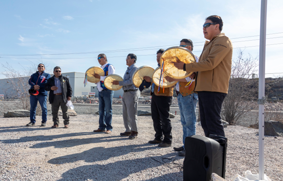 5 men with Dene drums perform a song in front of an industrial site, with 2 men standing solemnly observing.