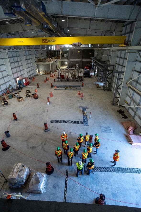 An aerial shot of a group of people in orange safety vests and green hard hats inside an industrial building.
