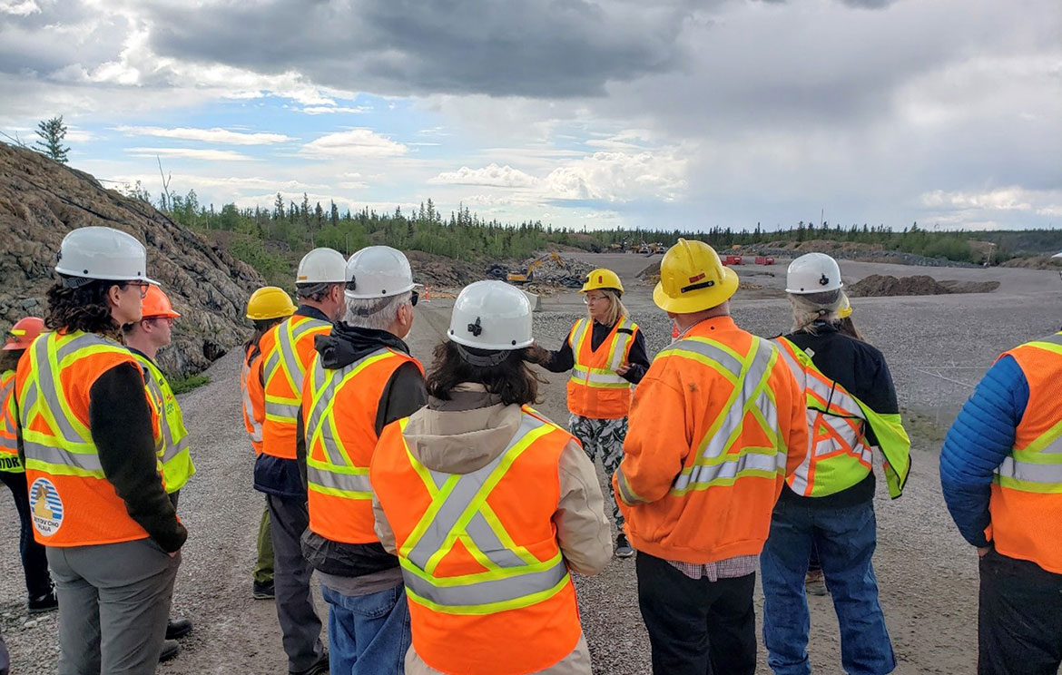 A group of people wearing safety vests and hard hats are shown standing on a gravel road with a rocky outcrop and trees visible in the background.