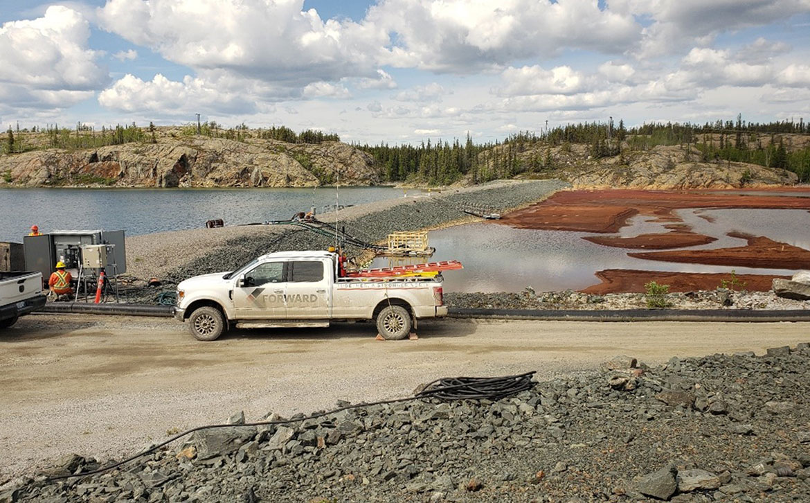 A white pickup truck is parked in front of 2 ponds separated by a gravel berm. The Polishing Pond is on the left and has clear water while the Settling Pond is on the right and is partially covered by ferric sulfate, the product used to treat the water, which is reddish in colour.