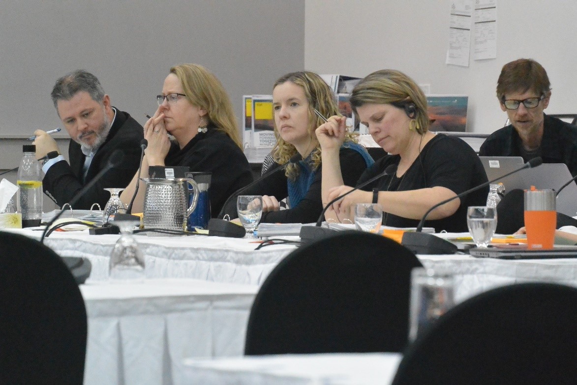 Five people are visible sitting behind tables, listening to a presentation.