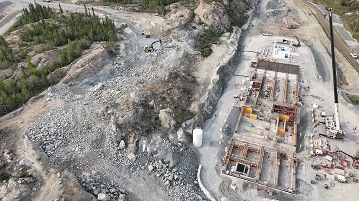 Vue aérienne d'un chantier de construction montrant les fondations d'un bâtiment sur la droite et des roches dynamitées par une excavatrice et un camion-benne sur la gauche.