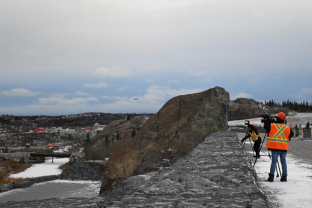 Two video journalists with video cameras and dressed in orange high-visibility safety vests and hard hats record footage from a rocky platform overlooking a mine site. 