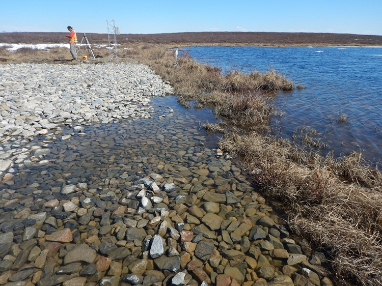 A worker in a safety vest stands the shore of Hambone Lake, with equipment used to measure water levels.