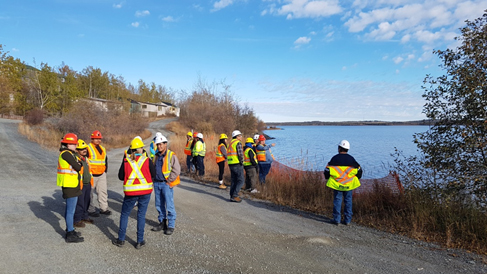 A group of people in personal protective equipment are seen in front of old, dilapidated houses with boarded up windows and a lake.