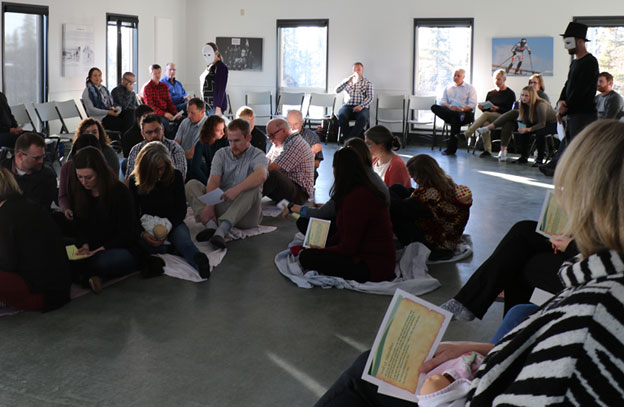 Cette photo montre des membres de l'équipe du projet participant à l'exercice de la couverture. Certains membres, représentant les peuples autochtones, sont assis par terre sur des couvertures qui représentent les terres autochtones, lesquelles ont été réduites et divisées tout au long de l'exercice. D'autres membres s'assoient en cercle autour des couvertures, car ils représentent des Autochtones qui sont morts et qui ont été retirés de l'exercice. Deux personnes masquées en blanc, qui représentent les colonisateurs, entourent les membres de l'équipe assis sur la couverture.