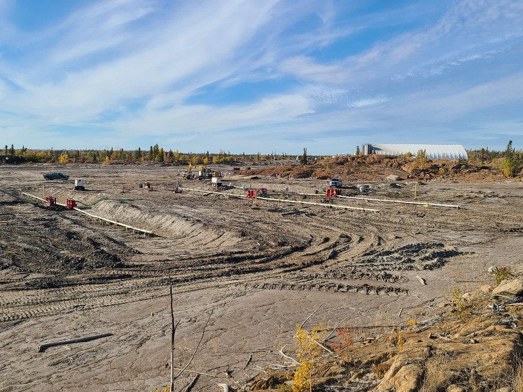 De-watering equipment is installed on a tailings pond, on which some trucks are located, with trees and a building visible in the background.