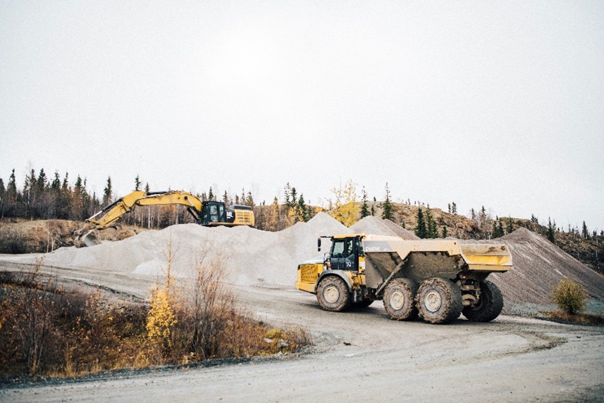 Un camion à benne vide roule sur une route non revêtue vers une excavatrice sur une grosse pile d’agrégats