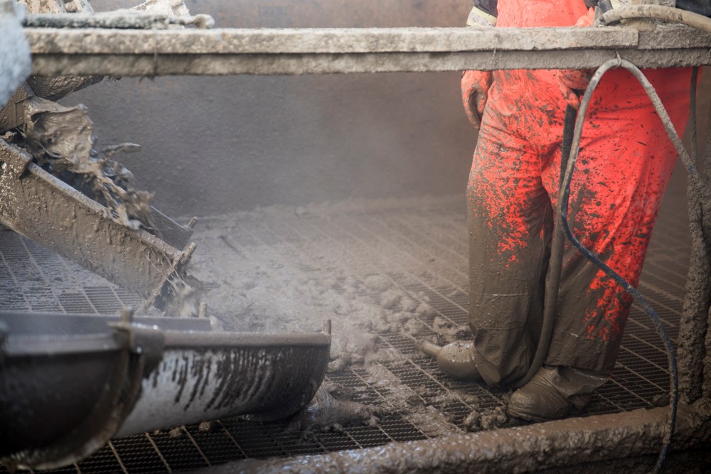 Troughs pour cemented paste backfill through a grate into the underground of the mine. The lower half of a worker wearing personal protective equipment is visible standing on the grate holding a hose in place with their boot.